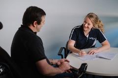 Indigo_OT_NDIS Male in black shirt in wheelchair at table with female smiling in Indigo uniform with pen and forms