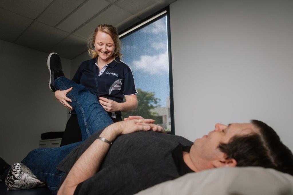 Female smiling wearing Indigo uniform stretching right leg of male lying down